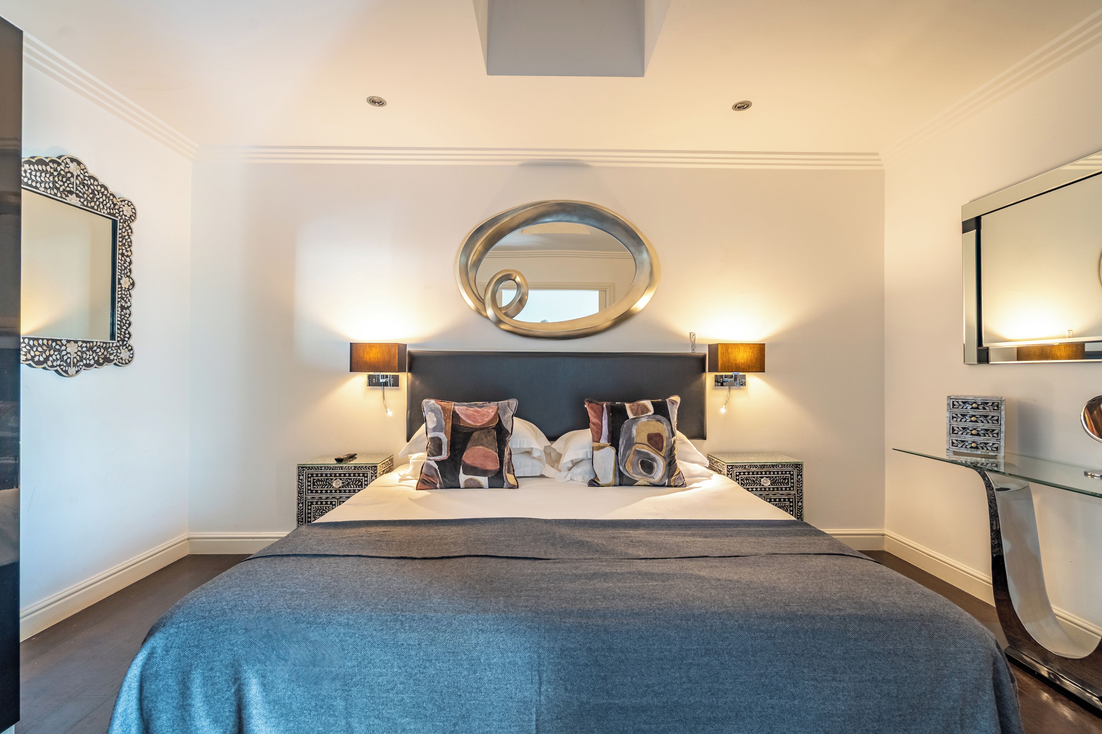 A modern bedroom with a large bed, decorative pillows, a round mirror above the bed, and two bedside tables with lamps.