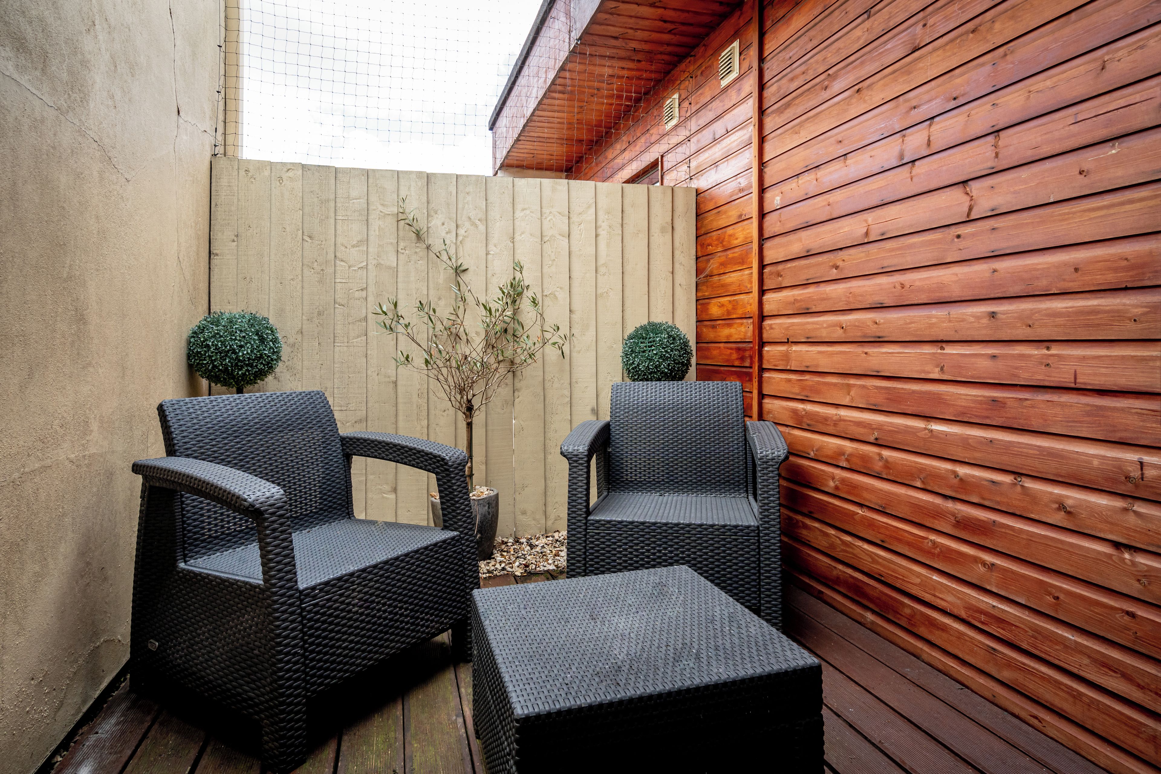 An outdoor seating area with two wicker chairs and a small table, surrounded by wooden walls and a concrete fence.