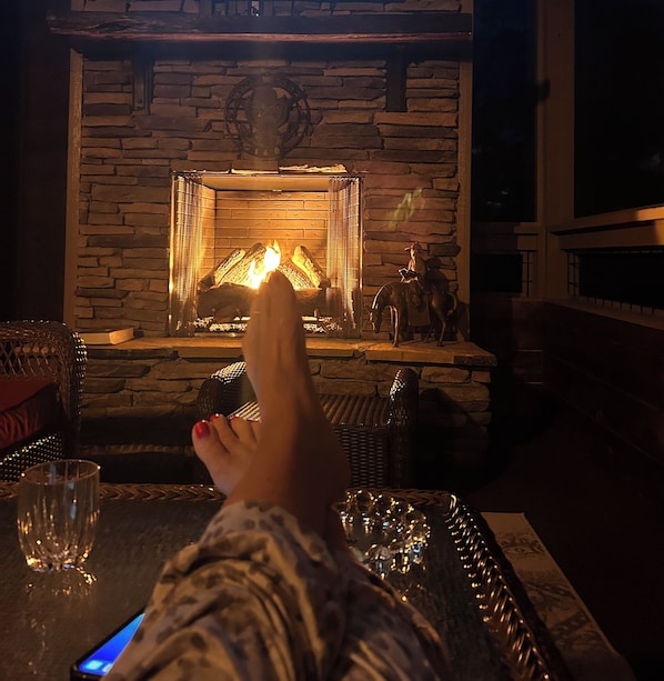 Une cheminée en pierre avec un feu qui crépite, une table en verre sur laquelle repose un verre, et les pieds d’une personne posés sur la table.