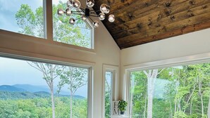 Un espace repas avec une table et des chaises en verre, de grandes fenêtres offrant une vue sur les arbres et les montagnes, et un plafond en bois.