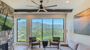 A living room with a stone fireplace, two armchairs, a sofa, a coffee table, and a large window offering a mountain view.