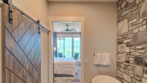 A modern bathroom with a stone accent wall, a freestanding bathtub, and a view of a bedroom with a bed and a ceiling fan.