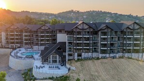 A multi-story hotel with a pool area, stone facade, and a view of a hilly landscape in the background.
