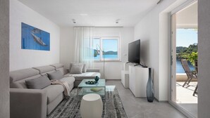 Ein modernes Wohnzimmer mit einer Couch, einem Couchtisch und einem Fernseher. Ein großes Fenster bietet Blick ins Freie.