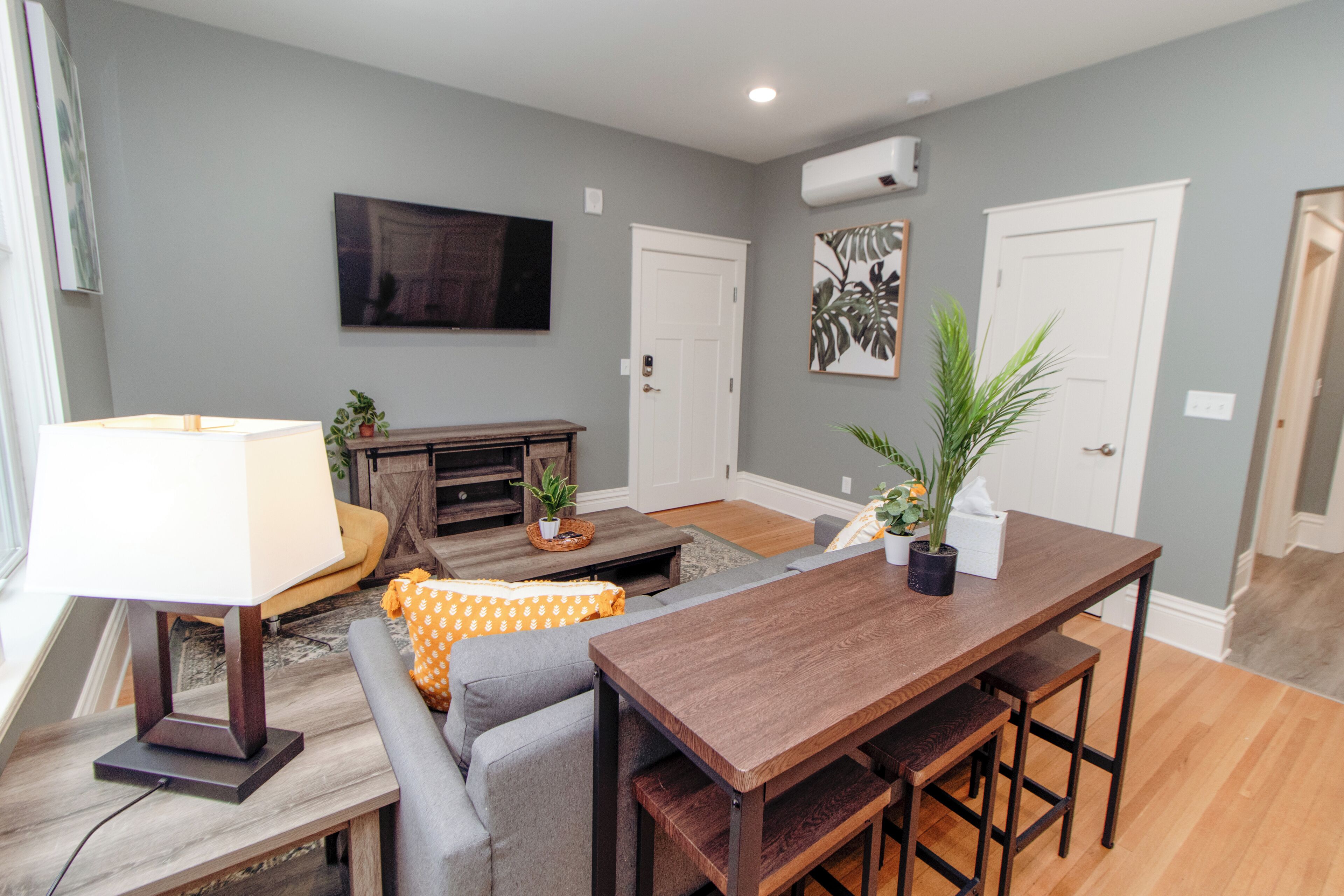 A modern living room with a wooden dining table, gray sofa, and a TV mounted on the wall.