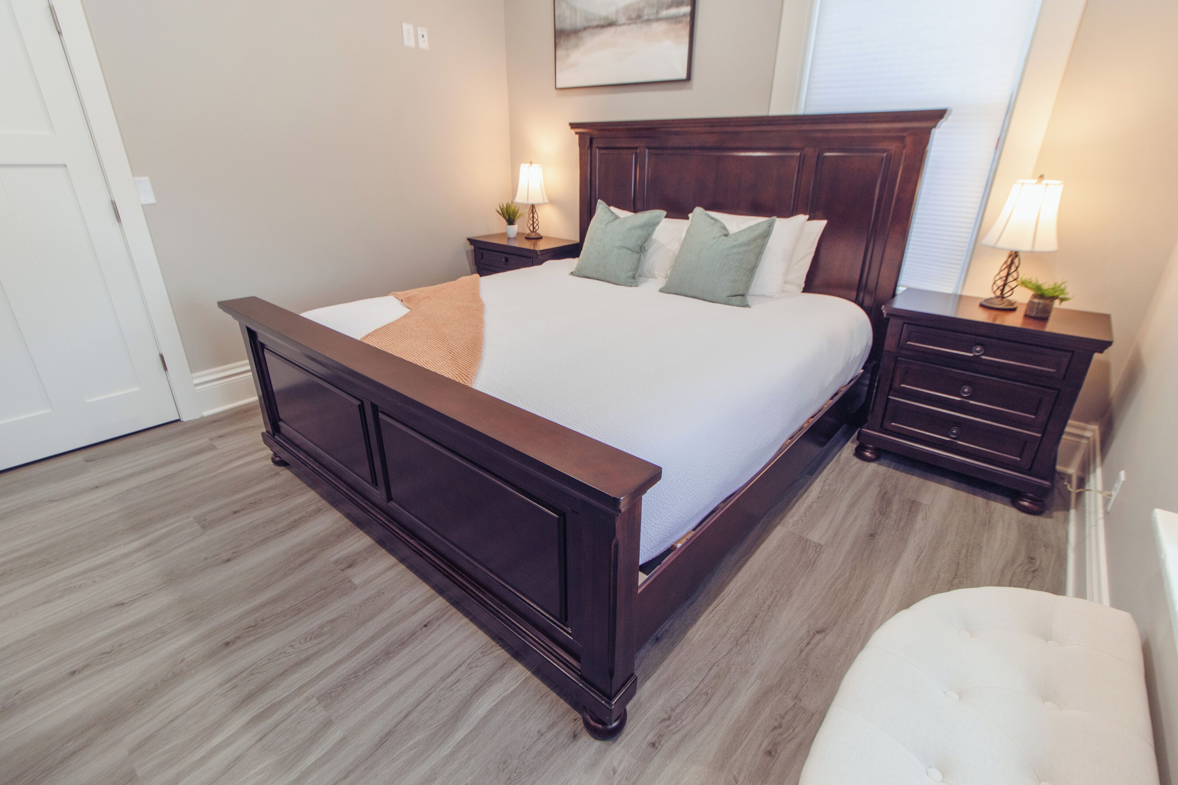 Une chambre à coucher avec un grand lit, une tête de lit en bois, deux tables de chevet avec des lampes, une petite chaise et une fenêtre avec des stores.