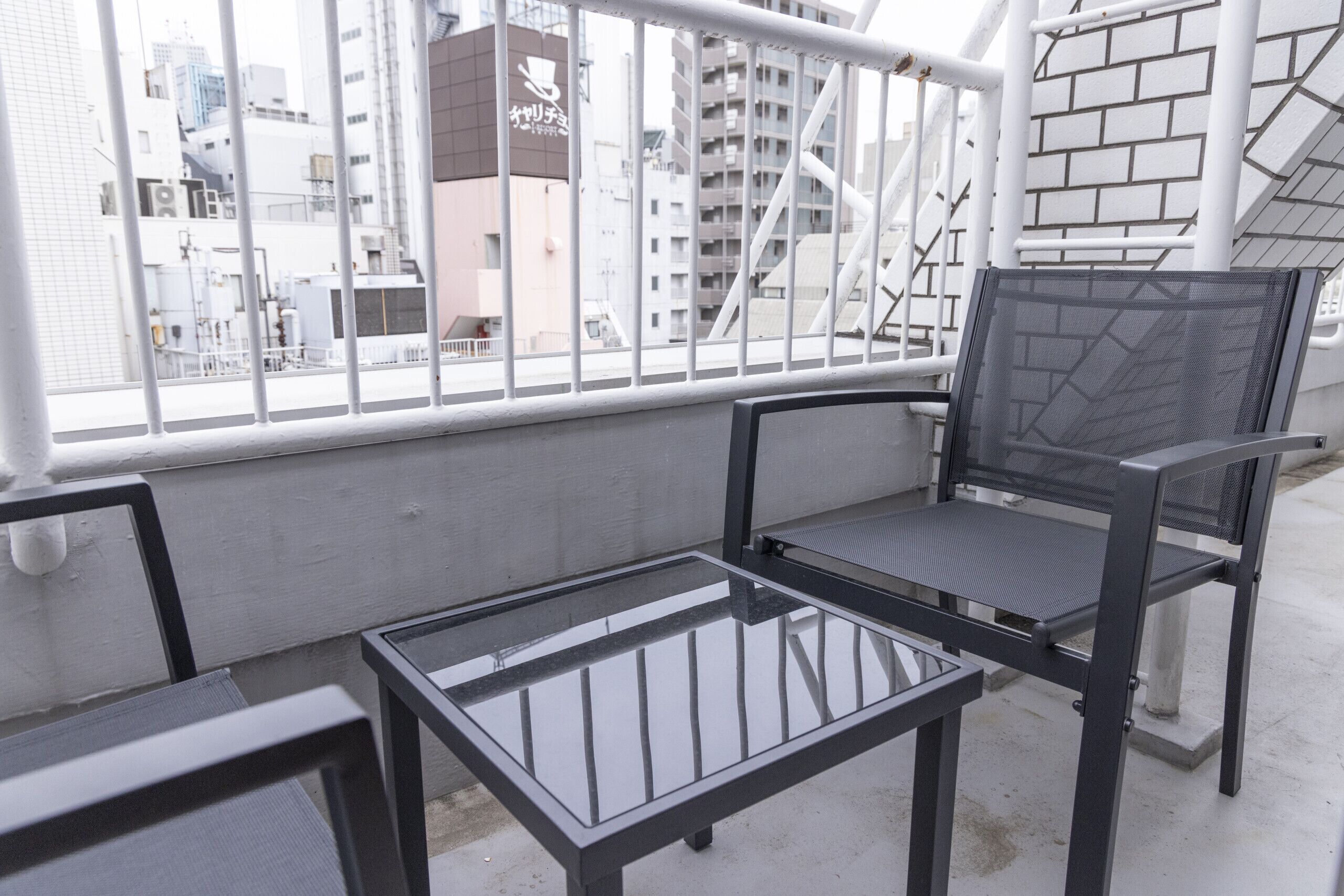 A balcony with a glass table and two chairs, overlooking a cityscape with buildings and a brick wall.