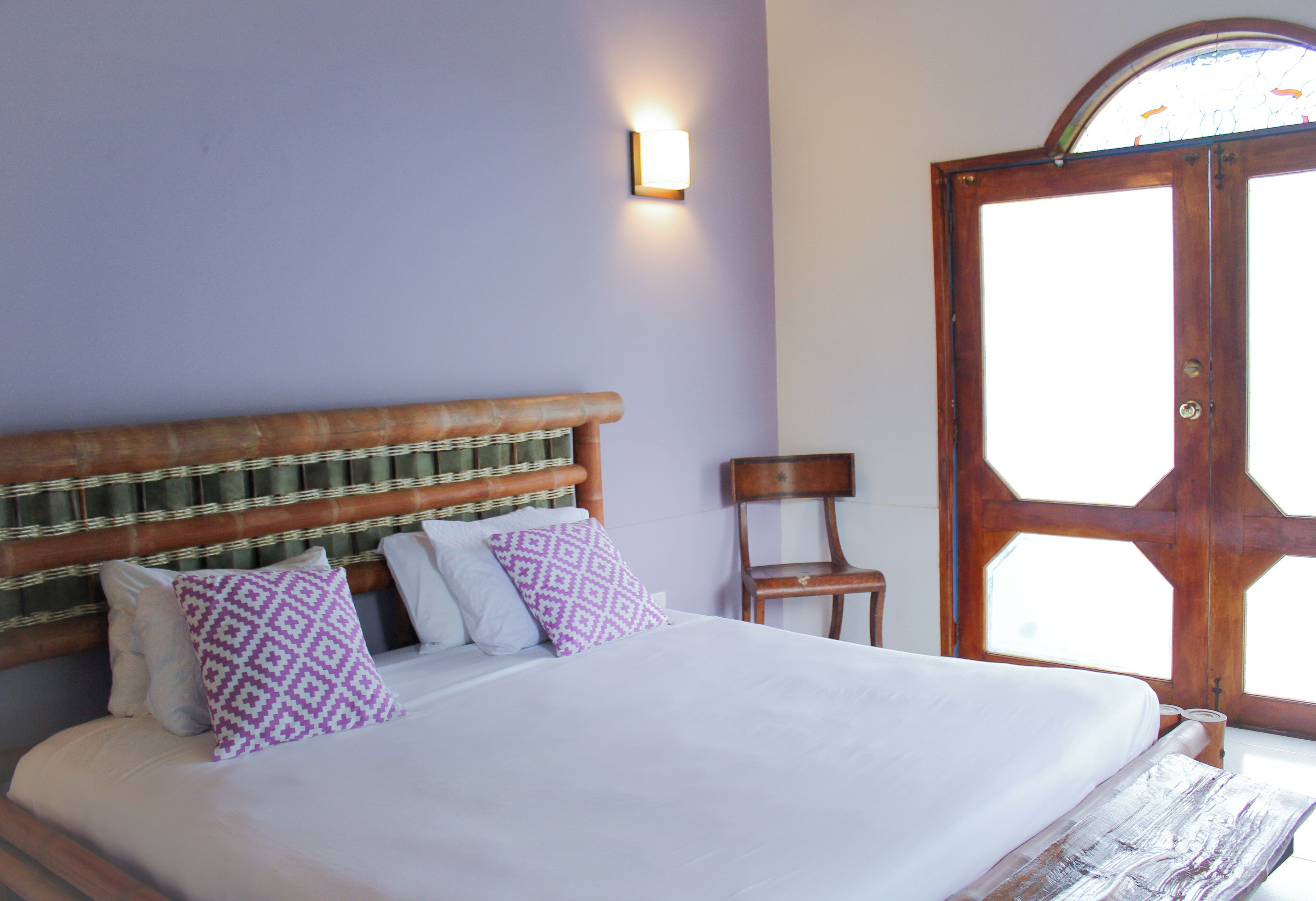A bedroom with a wooden headboard, a bed with white bedding, a chair, and a door with a stained glass window.