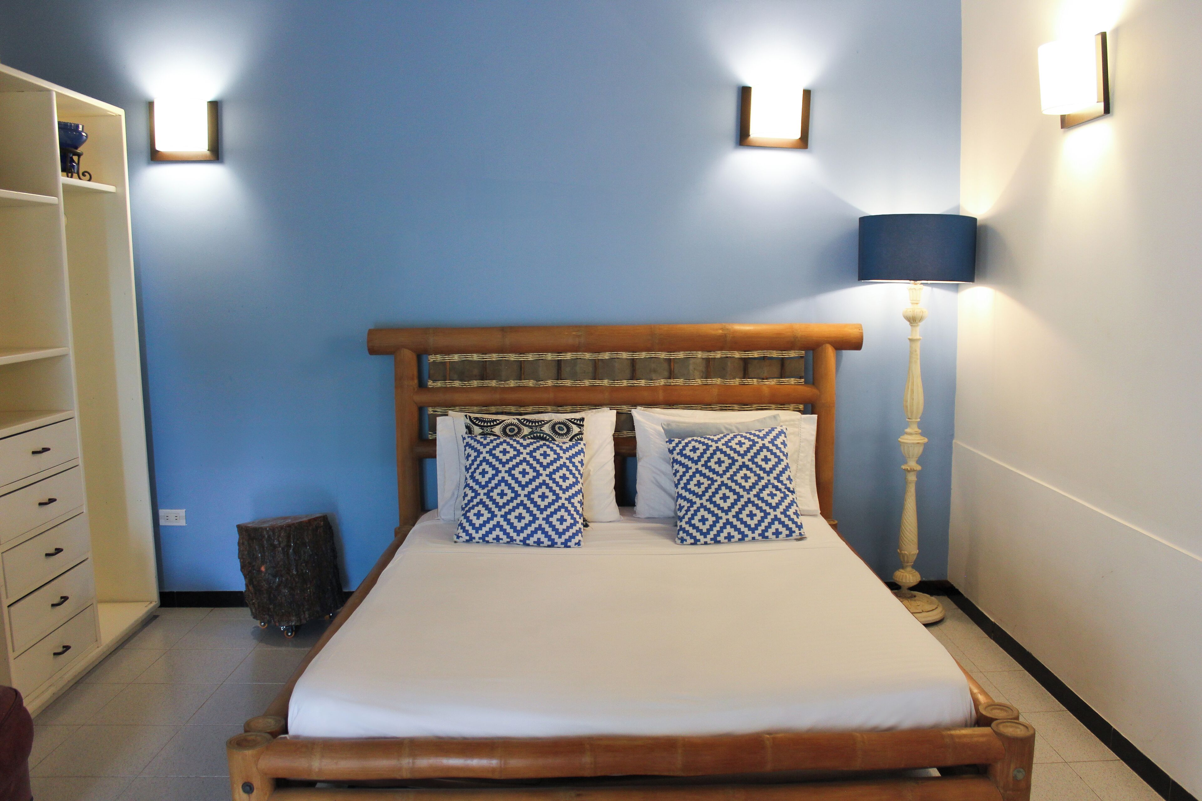 Un letto con testiera in legno, due cuscini decorati e una coperta bianca. Si nota una lampada da comodino e uno sgabello di legno.
