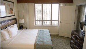 A hotel room with a large bed, a nightstand with a lamp, a dresser, and a window with a view of high-rise buildings. - Hilton Hawaiian Village Lagoon Tower 2BR 2BA sleeps 6 (Honolulu)