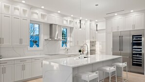 A modern kitchen with white cabinetry, a large island with bar stools, stainless steel appliances, and a window with a view of trees. - Brand new 4bdr, 5 bath luxury house 1 mile from 5th Avenue and Beached (Naples)