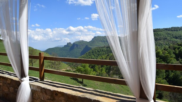 A balcony with white curtains, a wooden railing, and a view of green hills and a clear blue sky. - Enjoy nature in its purest form (Vallfogona de Ripollès)