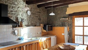 A rustic kitchen with exposed beams, a stone wall, a wooden table, and a window with a view. - Enjoy nature in its purest form (Vallfogona de Ripollès)