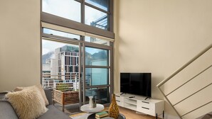 A modern living room with a grey sofa, a TV on a white stand, and a large window offering a view of a building and mountains. - Elegant 1 Bedroom Loft - Soho on Rose (Cape Town)