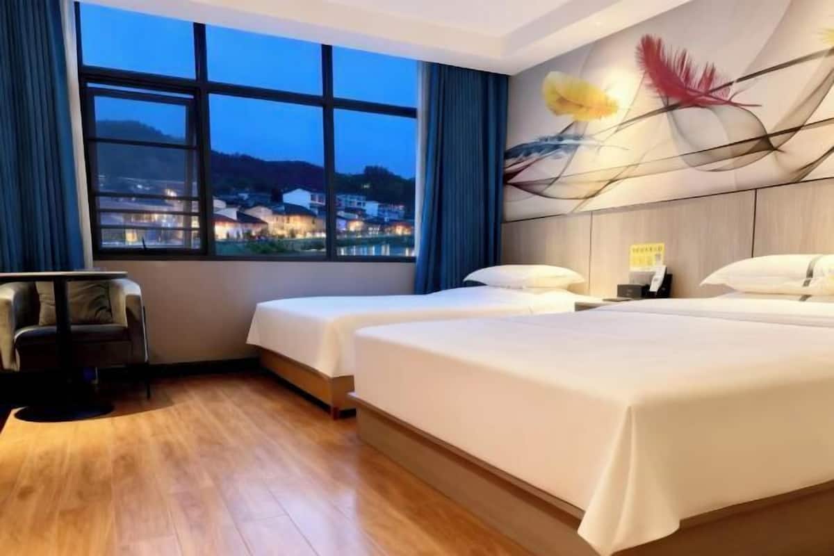 A hotel room with two beds, a chair, a large window with a view of buildings, and a decorative wall piece.