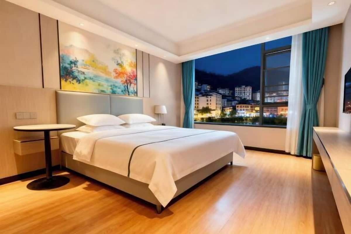 A modern hotel room with a large bed, a small round table, a painting on the wall, and a view of a cityscape at night.