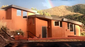 Una moderna casa a due piani con tetto piatto e ampie finestre, sullo sfondo di montagne e con un arcobaleno visibile.