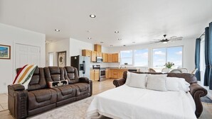 Un salón espacioso con un amplio sofá de cuero marrón, una cama blanca y una cocina al fondo.