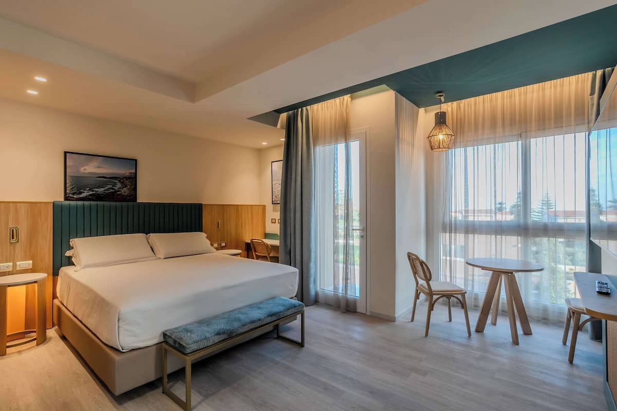 A modern hotel room with a large bed, a bench, a dining area with a table and chairs, and a view of the outdoors through large windows.