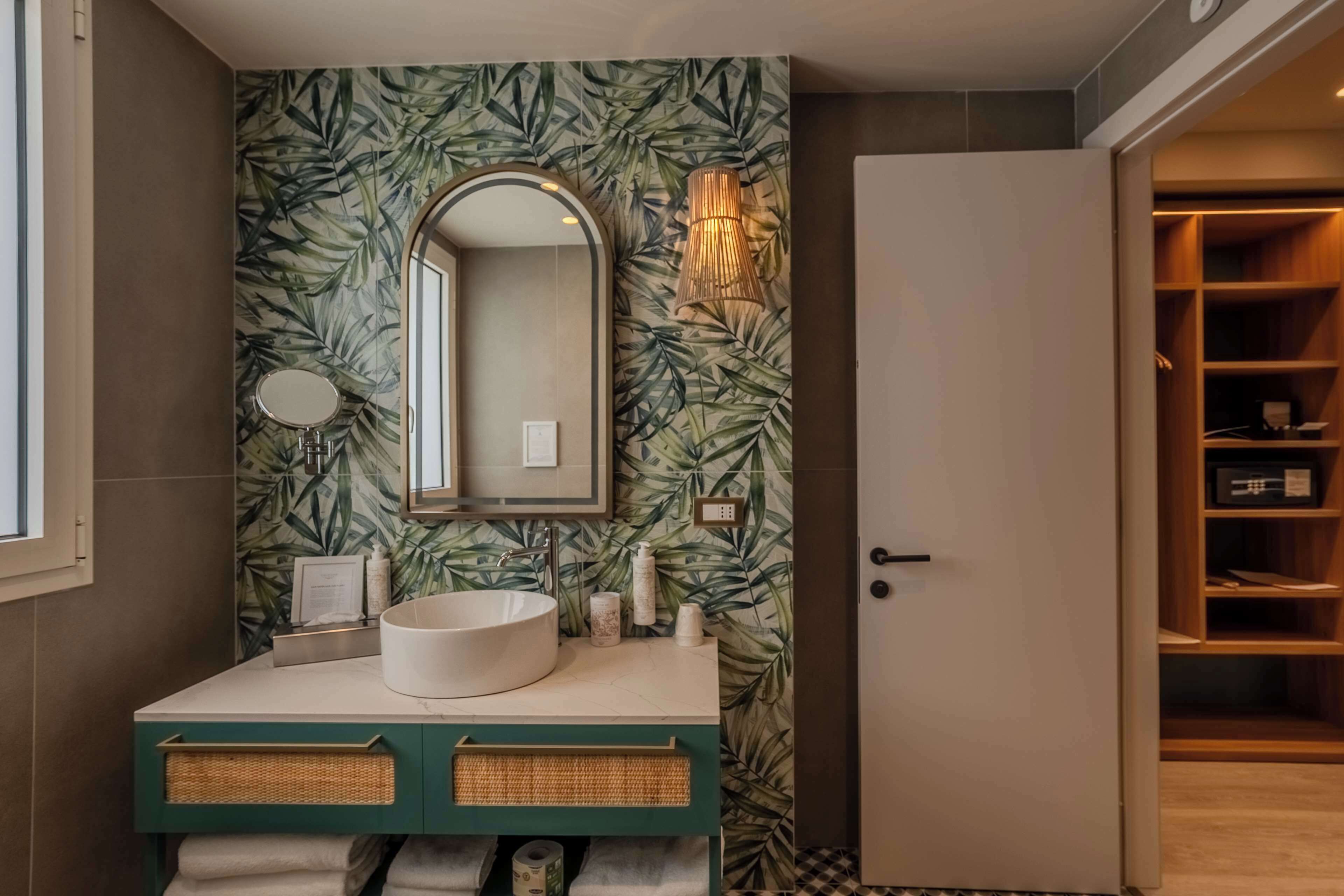 A bathroom with a round sink, a mirror, and a wall with a leafy pattern. There is a door to a closet and a shelf with toiletries.