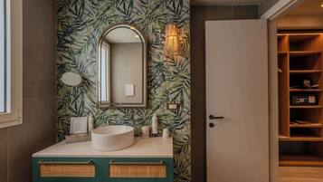 A bathroom with a round sink, a mirror, and a wall with a leafy pattern. There is a door to a closet and a shelf with toiletries.