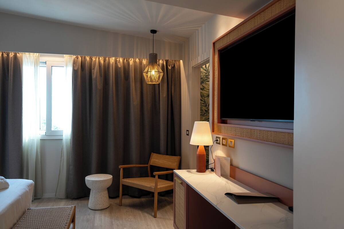 A hotel room with a bed, a chair, a small table, a lamp, a television, and a window with curtains.