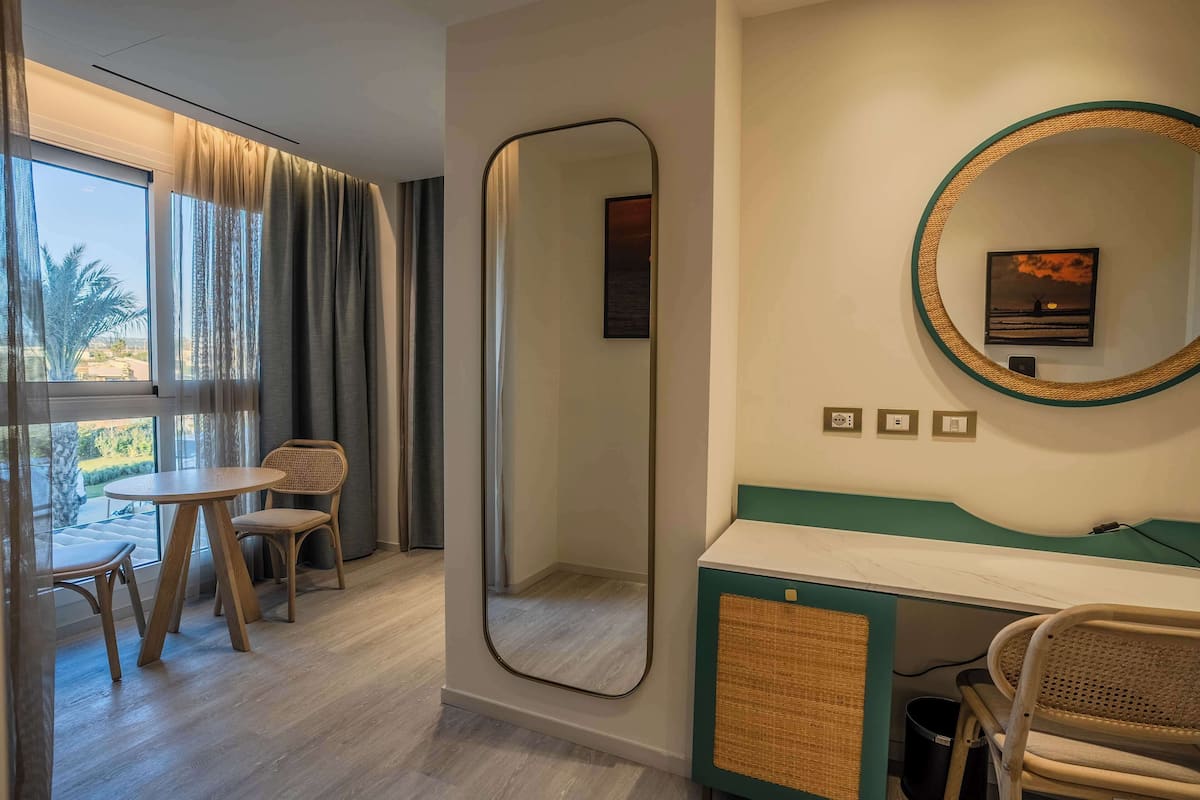 A hotel room with a large window, a round mirror, a round table with two chairs, a small round table, and a green accent wall.
