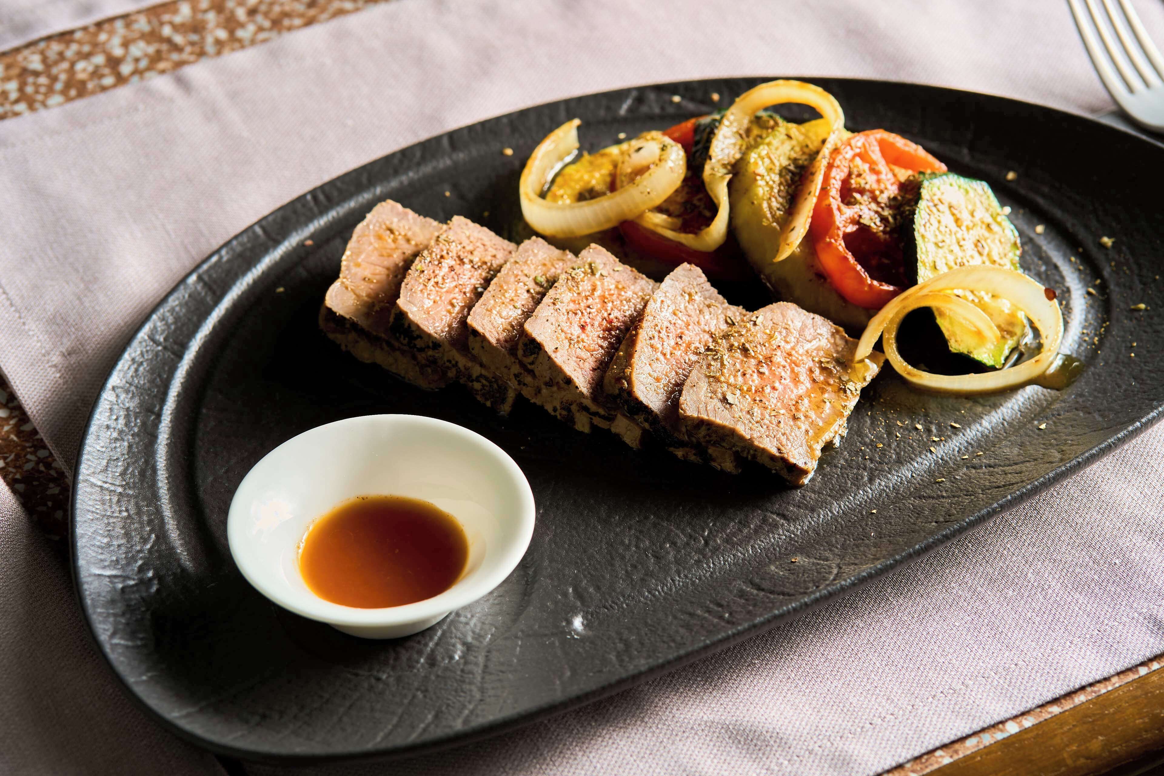 A dish with sliced meat and vegetables on a black plate, accompanied by a small bowl of sauce.