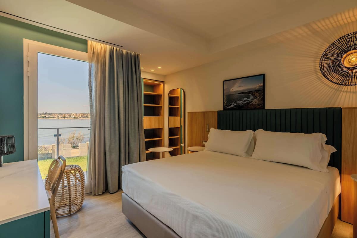 A hotel room with a large bed, a desk, a chair, a round wicker side table, a view of the outdoors, and built-in shelving.