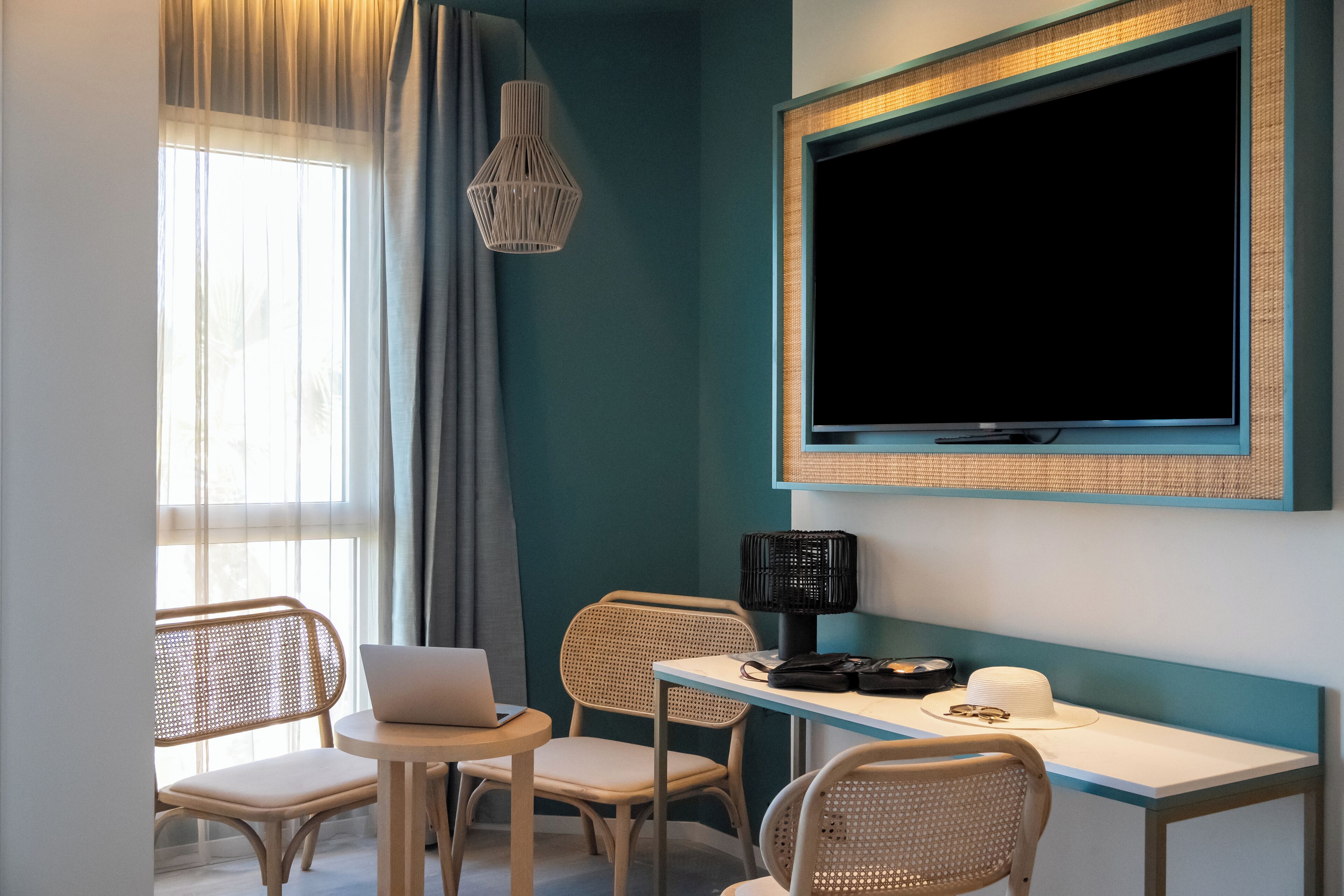 A modern room with teal walls, a large TV mounted on the wall, a laptop on a small table, wicker chairs, and a woven pendant light.
