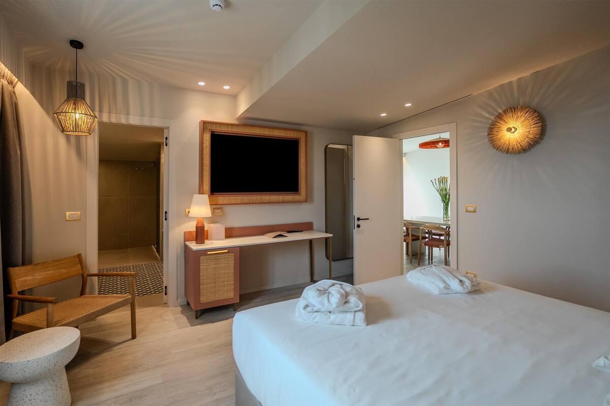 A hotel room with a bed, a desk, a chair, a mirror, and a hanging light fixture.