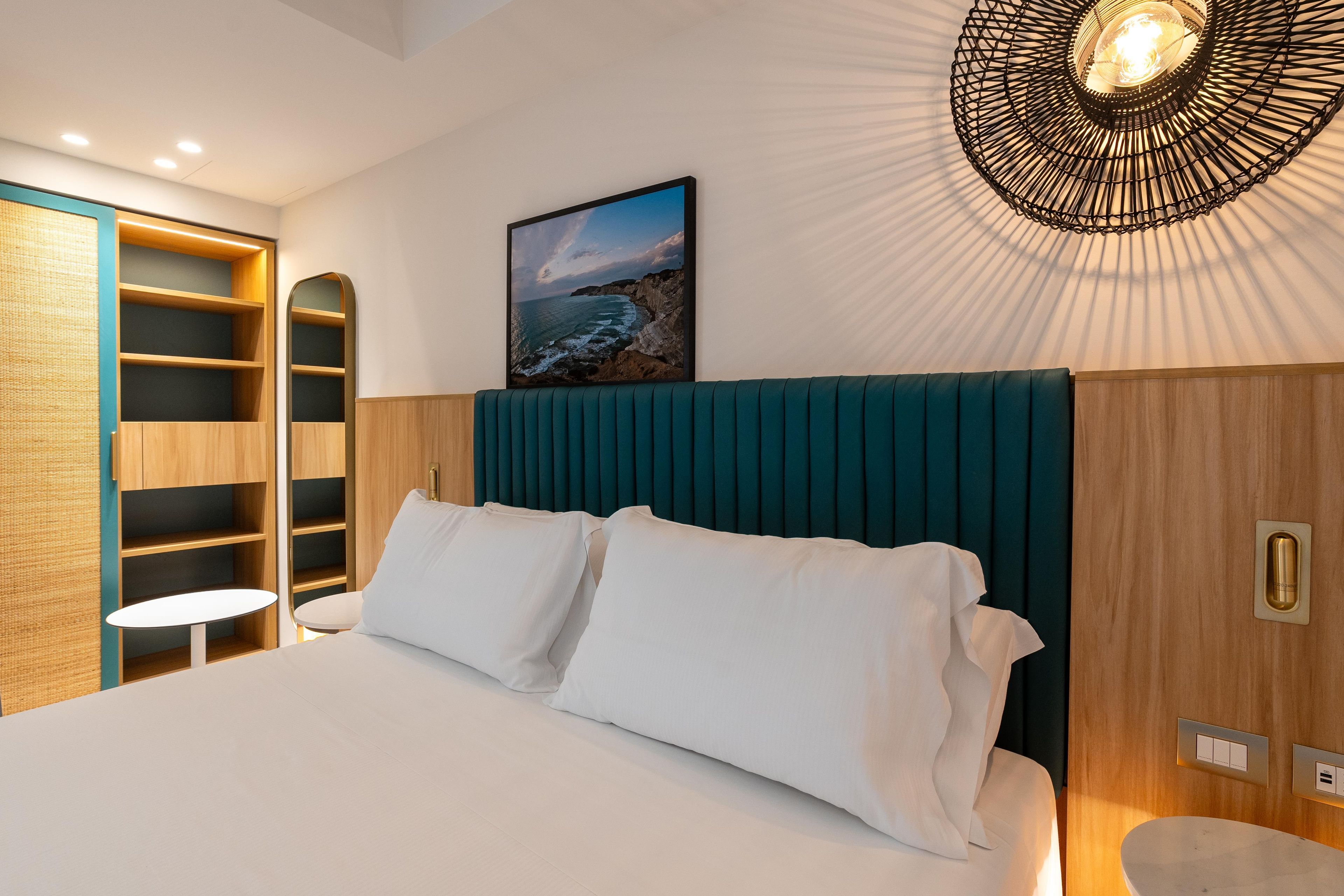 A hotel room with a bed, a view of the sea, a wooden wardrobe, and a unique ceiling light fixture.
