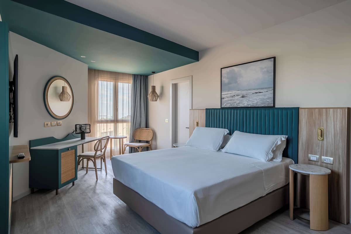 A hotel room with a large bed, a desk with a chair, a small table, and a view of the sea.