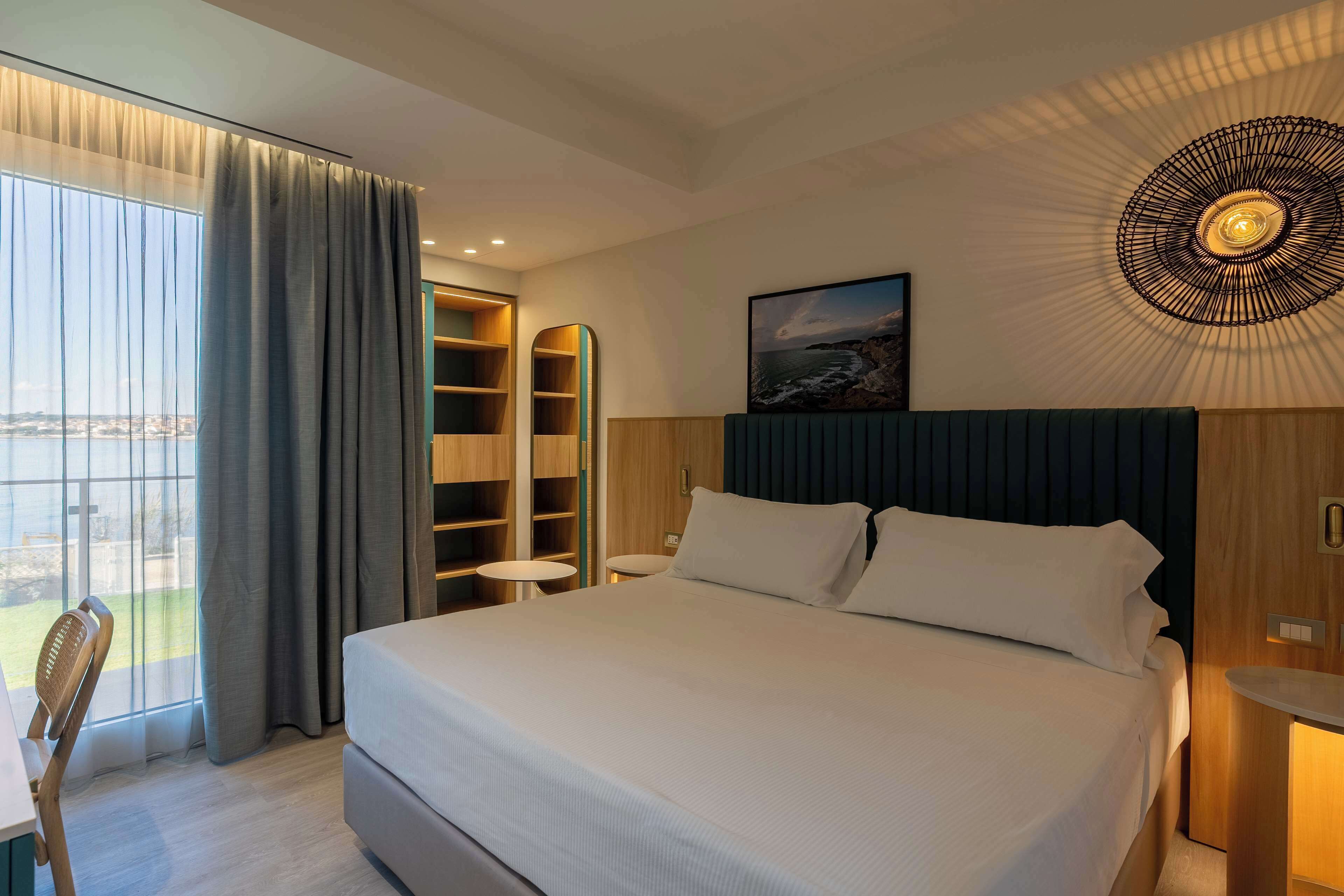 Une chambre d’hôtel dotée d’un grand lit, offrant une vue sur l’extérieur, d’une bibliothèque intégrée et d’un luminaire au plafond original.