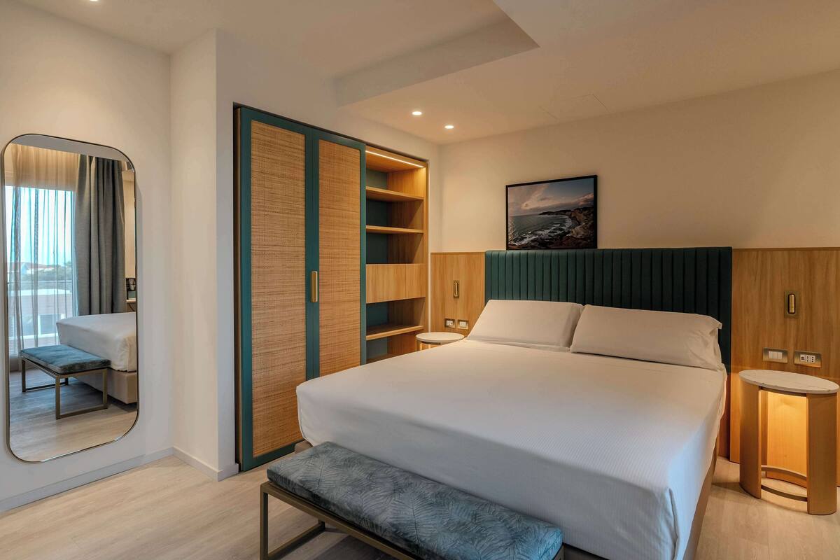 A modern hotel room with a large bed, a bench, a mirror, and built-in shelves.