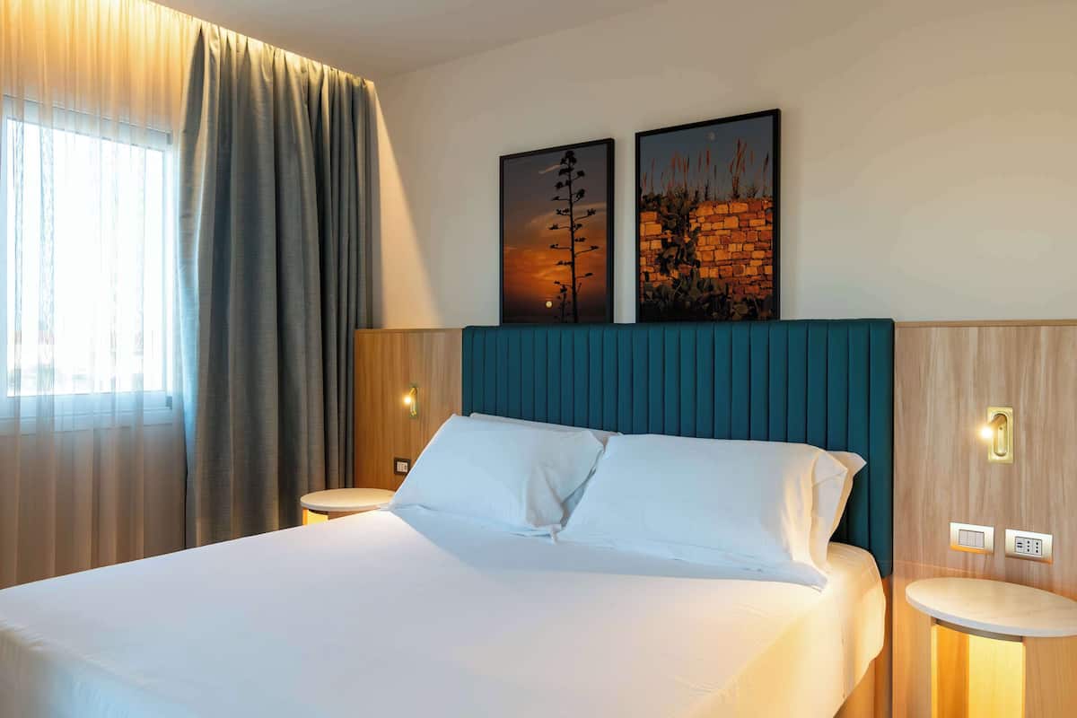 A hotel room with a large bed, two wall-mounted paintings, a wooden headboard, a bedside table, and a nightstand.