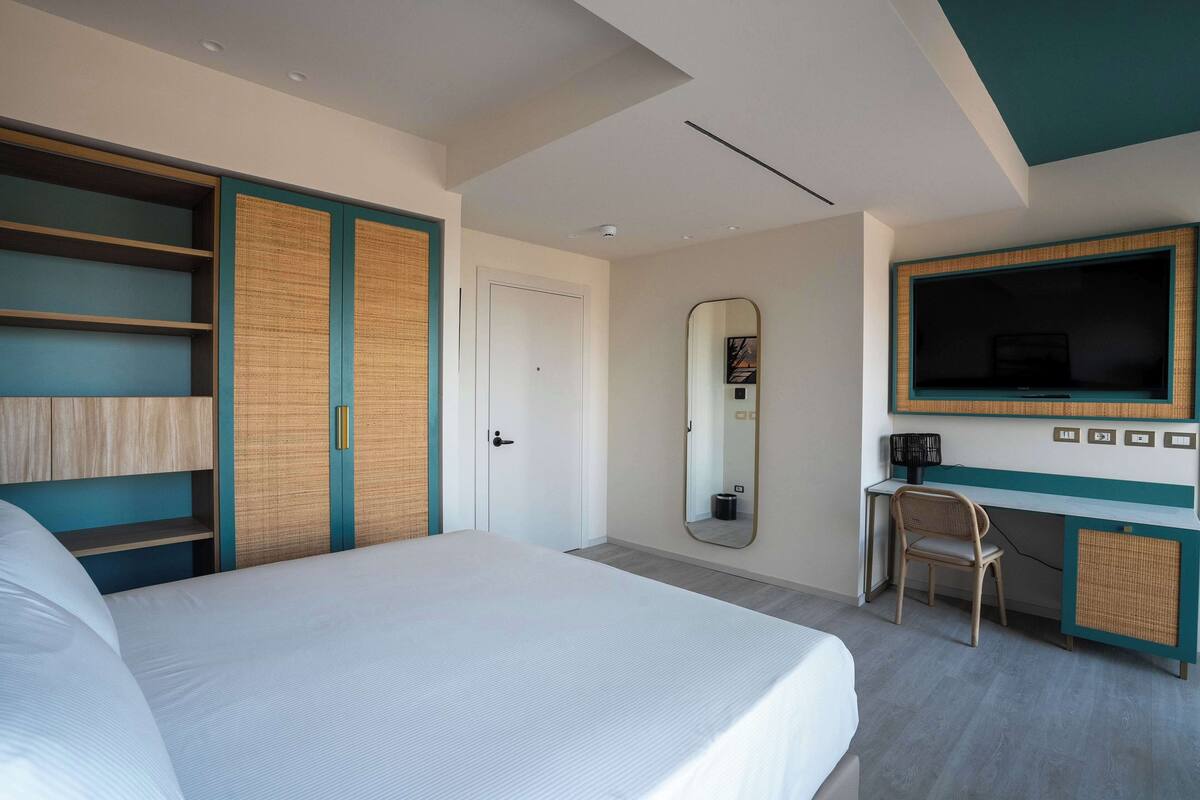 A modern hotel room with a large bed, a desk with a chair, a television, and a wardrobe with a mirror.