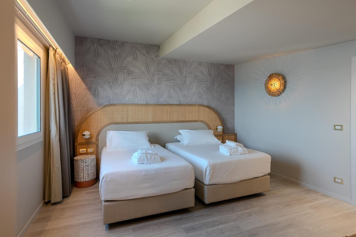 A hotel room with two beds, a patterned wall, a round wall light, and a window with curtains.