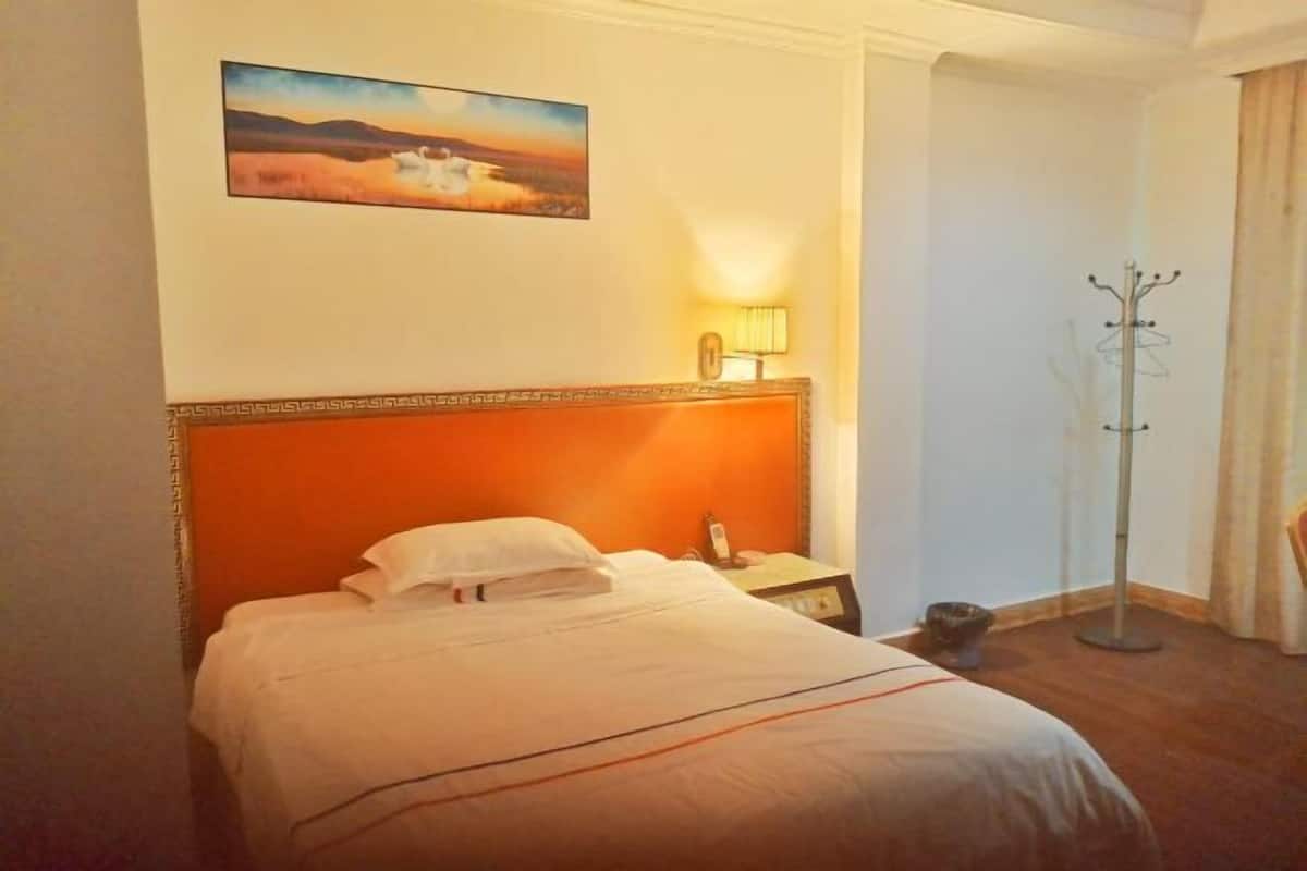 A hotel room with a bed, a nightstand, a lamp, a coat rack, and a painting on the wall.