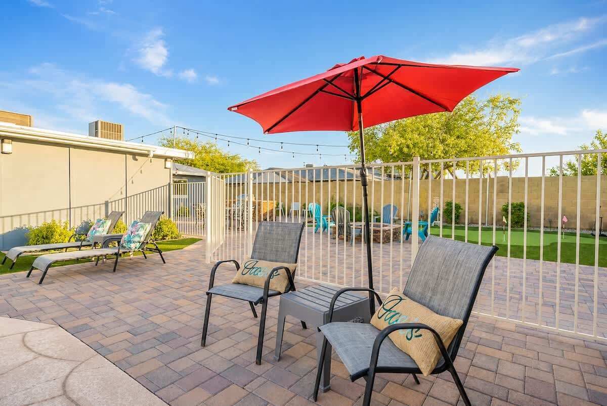 An outdoor patio with two lounge chairs and a red umbrella, surrounded by a white fence and a lawn.
