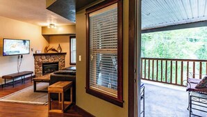 A living room with a stone fireplace, wooden furniture, and a large window with blinds. - Pool, Hot Tub & Gym | Near Fernie Alpine Resort (Fernie)