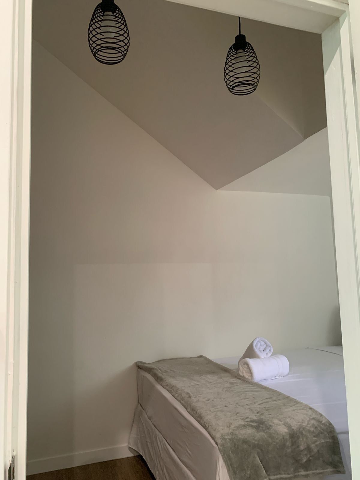 Une petite chambre avec un lit simple, recouverte d’une courtepointe grise et agrémentée d’une serviette blanche enroulée. Deux lampes suspendues noires sont fixées au plafond.