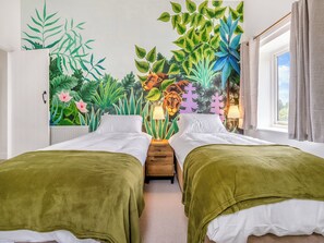 Et værelse med to senge, et vægmaleri med tropiske planter, et vindue med gardiner og en lampe på natbordet.