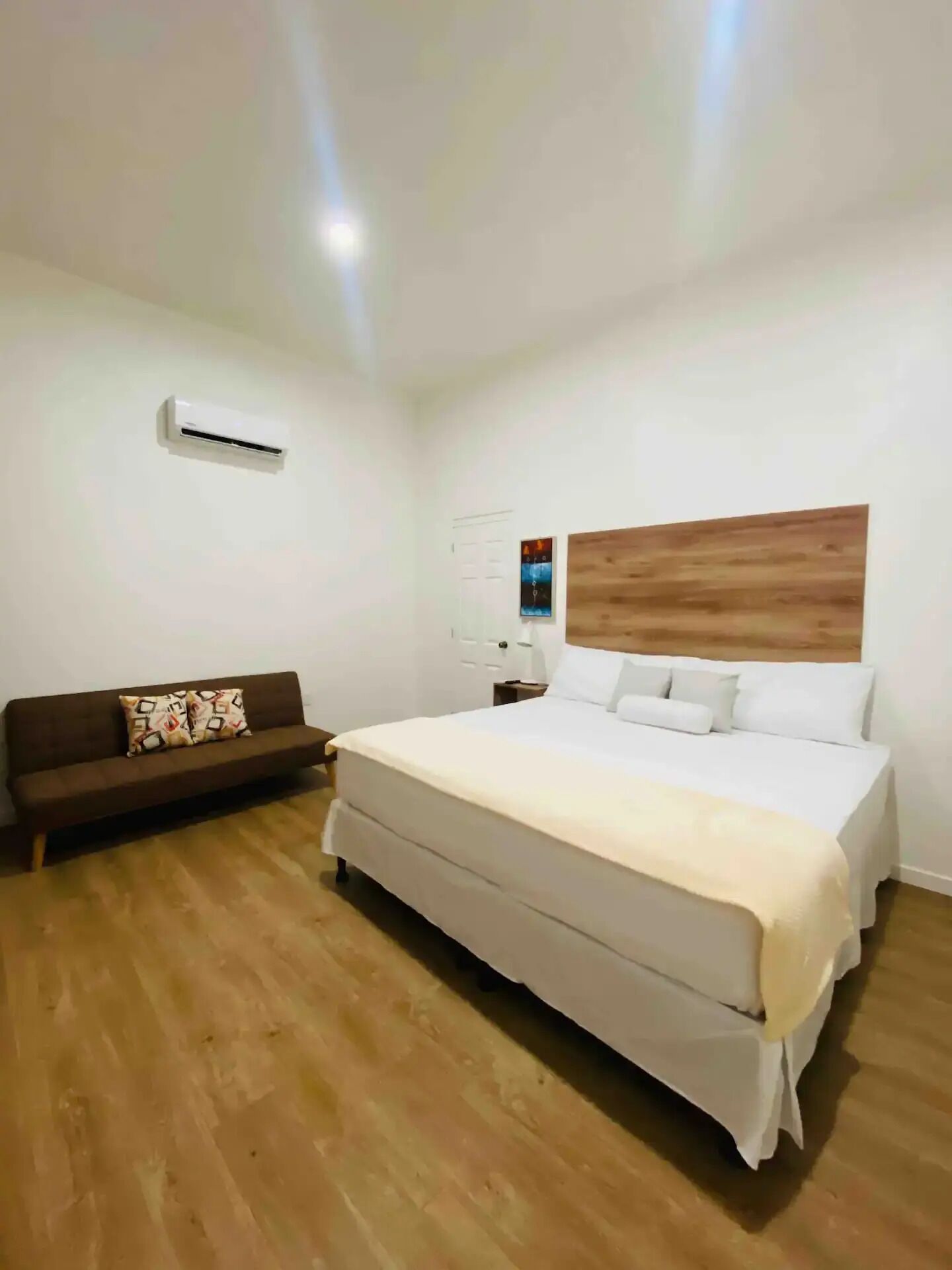 A bedroom with a large bed, a wooden headboard, a brown sofa, and an air conditioning unit on the wall.