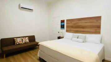 A bedroom with a large bed, a wooden headboard, a brown sofa, and an air conditioning unit on the wall.
