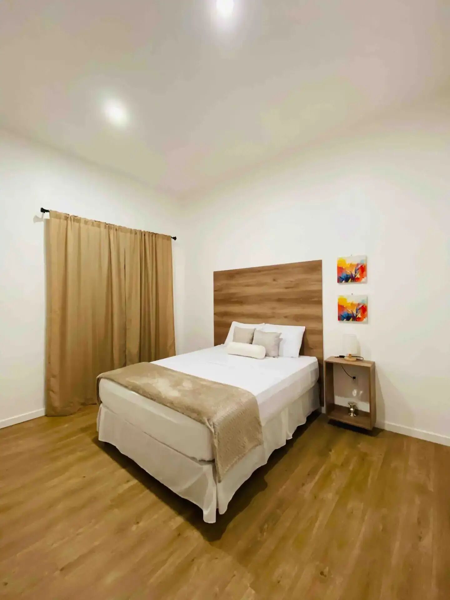 A neatly arranged bedroom with a wooden headboard, a bed with white bedding, a bedside table with a lamp, and a large window with curtains.