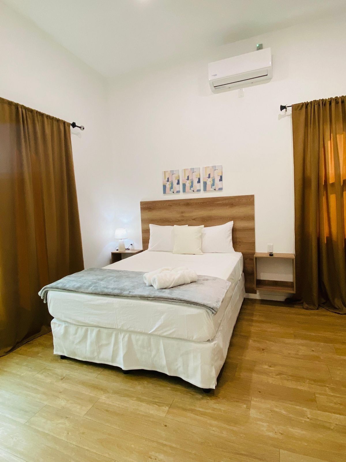 Een netjes opgemaakt bed met een houten hoofdbord, een nachtkastje met een lamp, en een wandairconditioner.