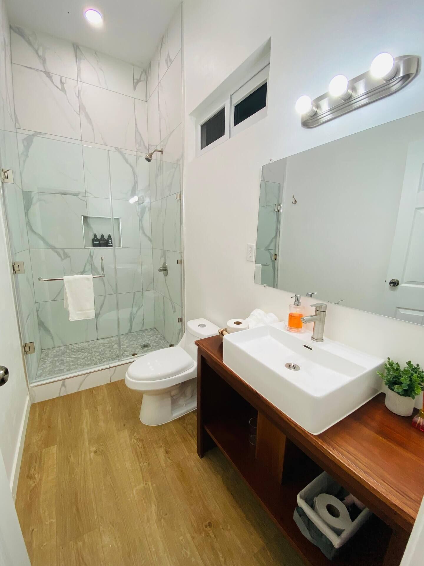 A modern bathroom with a glass-enclosed shower, a white toilet, a sink with a mirror above, and a small window.