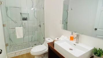 A modern bathroom with a glass-enclosed shower, a white toilet, a sink with a mirror above, and a small window.