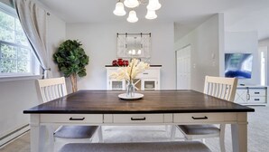 Une salle à manger avec une table en bois foncé, des chaises blanches, un lustre et une vue sur la cuisine et le salon.